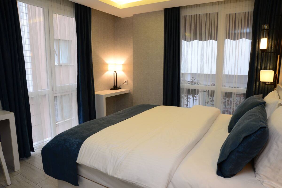 A hotel room with a large bed, a bedside table with a lamp, and a view through sheer curtains.