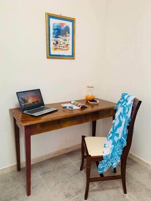 A wooden desk with a laptop, a glass pitcher of orange juice, and a book. A chair with a blue and white patterned cloth is positioned in front of the desk. - Agriturismo Trappeto Vecchio (San Demetrio Corone)