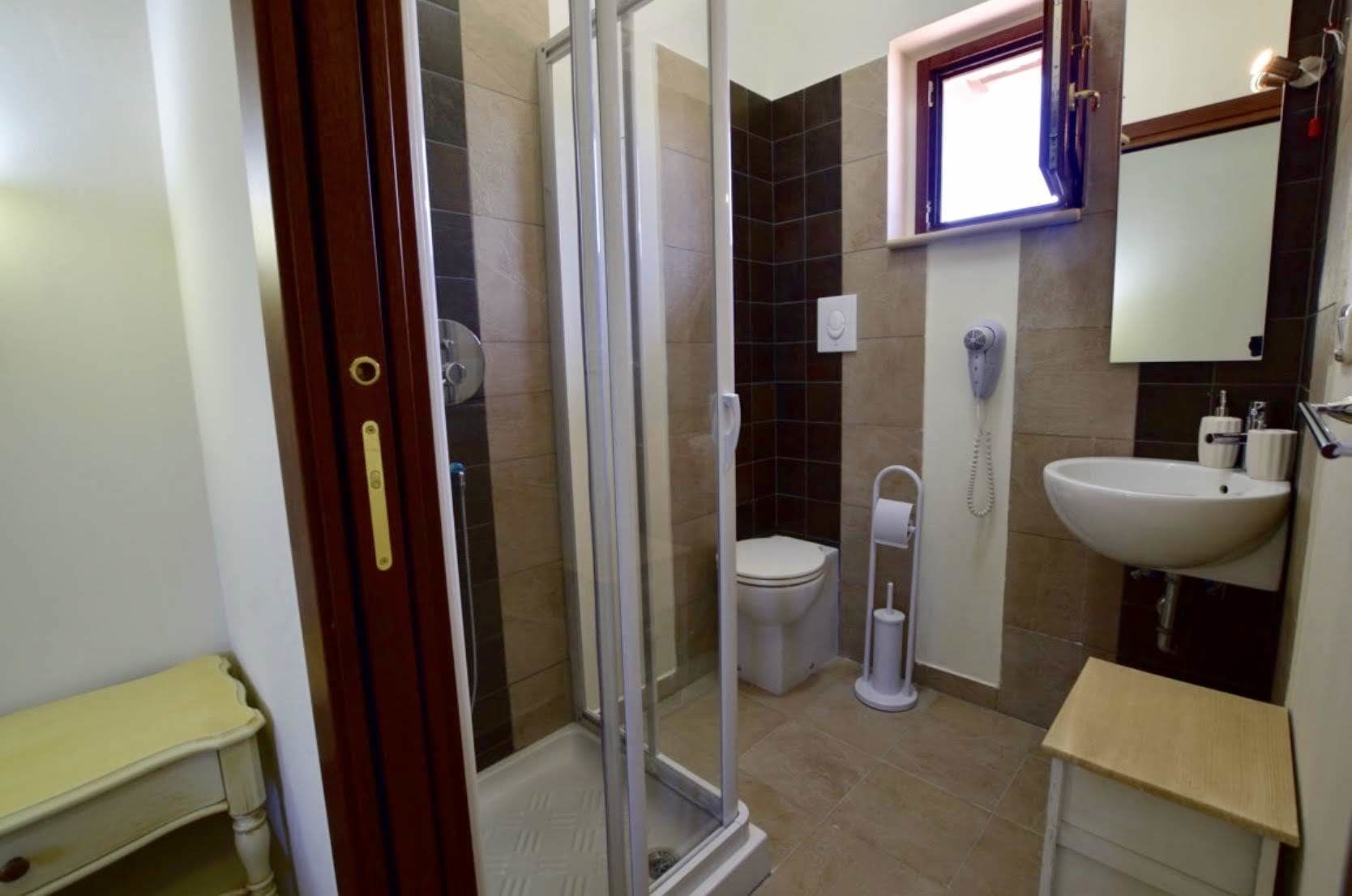 Bagno con box doccia in vetro, un water, un lavandino e una piccola finestra.