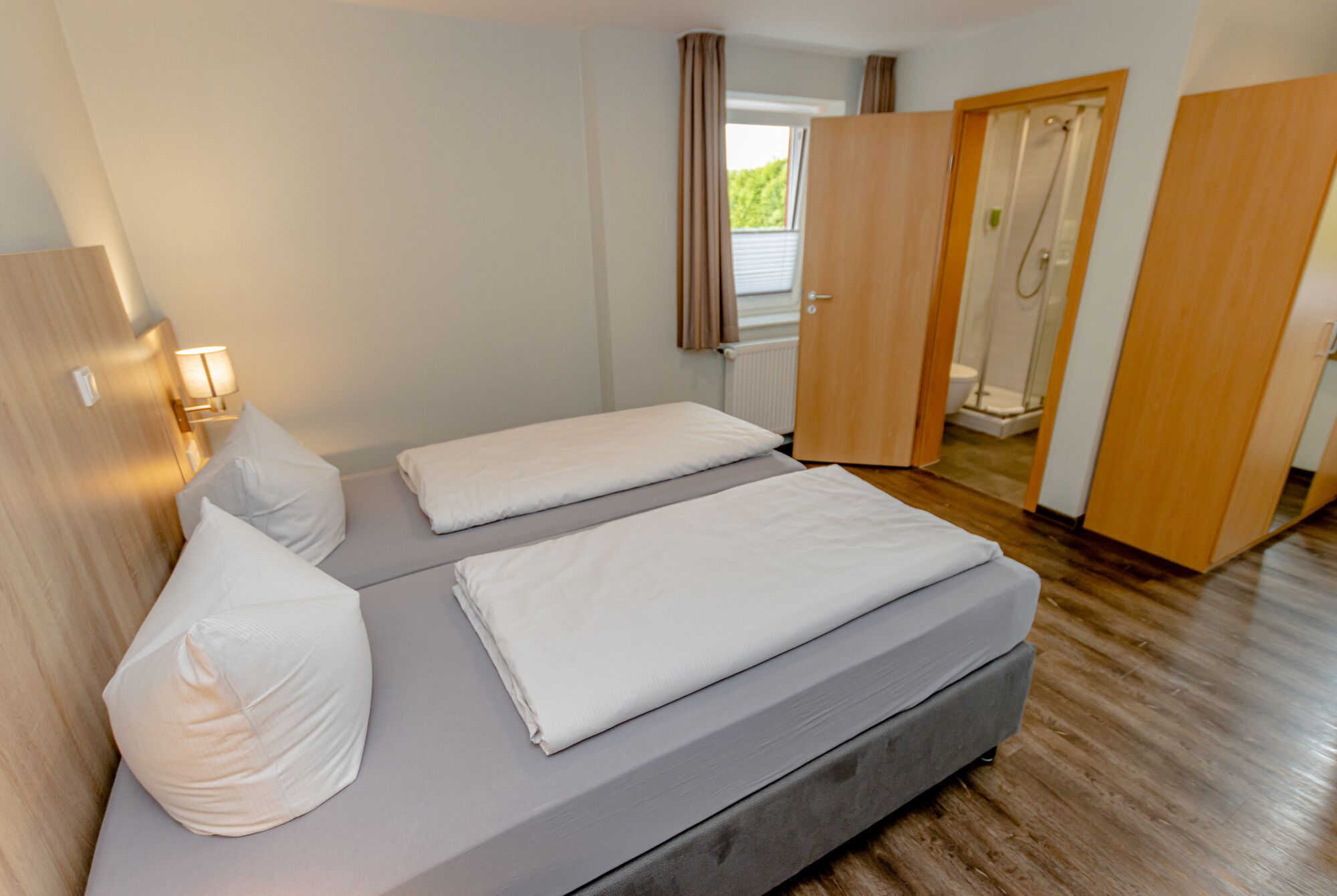 A bedroom with a bed, pillows, a nightstand, a lamp, and a view of a bathroom with a shower.