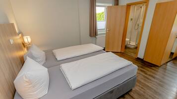 A bedroom with a bed, pillows, a nightstand, a lamp, and a view of a bathroom with a shower.