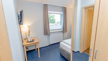 Una habitación de hotel pequeña y limpia, con una cama, una mesita de noche con lámpara, una ventana con cortinas y una puerta que da a otra habitación.