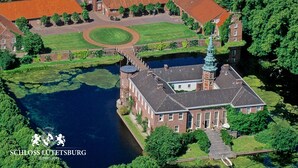 An aerial view of Schloss Lütetsburg, featuring a large brick building with a tower, surrounded by lush greenery and a body of water. - Hotel Herbers & RestaurantLeon (Sudbrookmerland)