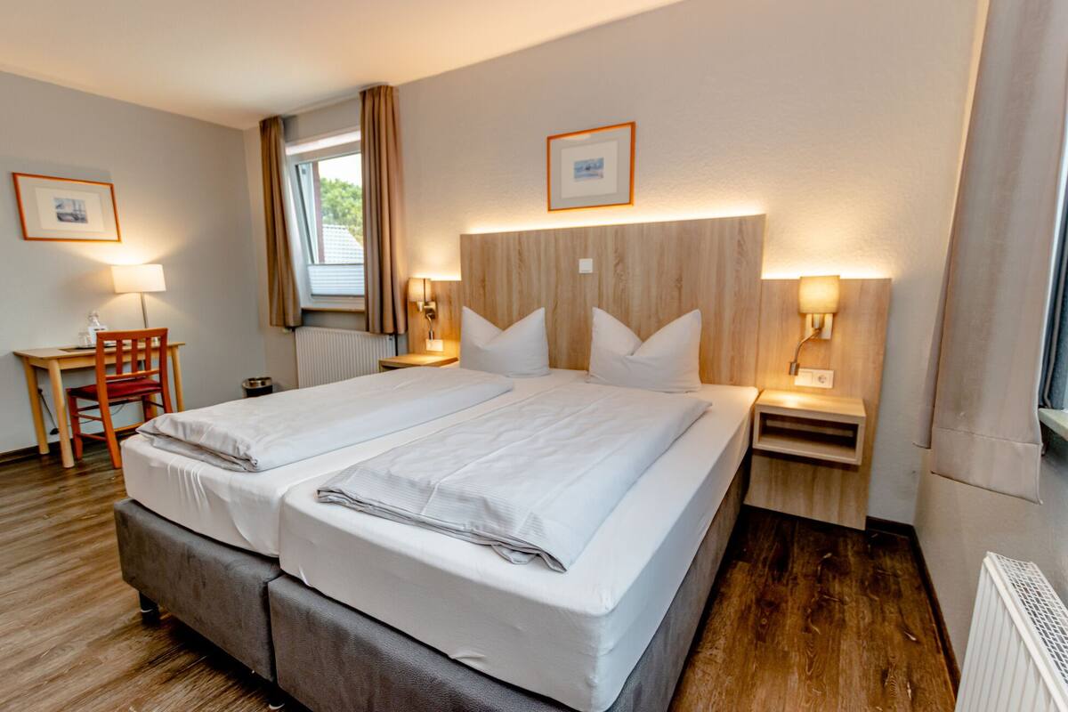 Een tweepersoonsbed met houten hoofdbord, twee nachtkastjes met lampen, een bureau met stoel, en een raam met gordijnen.