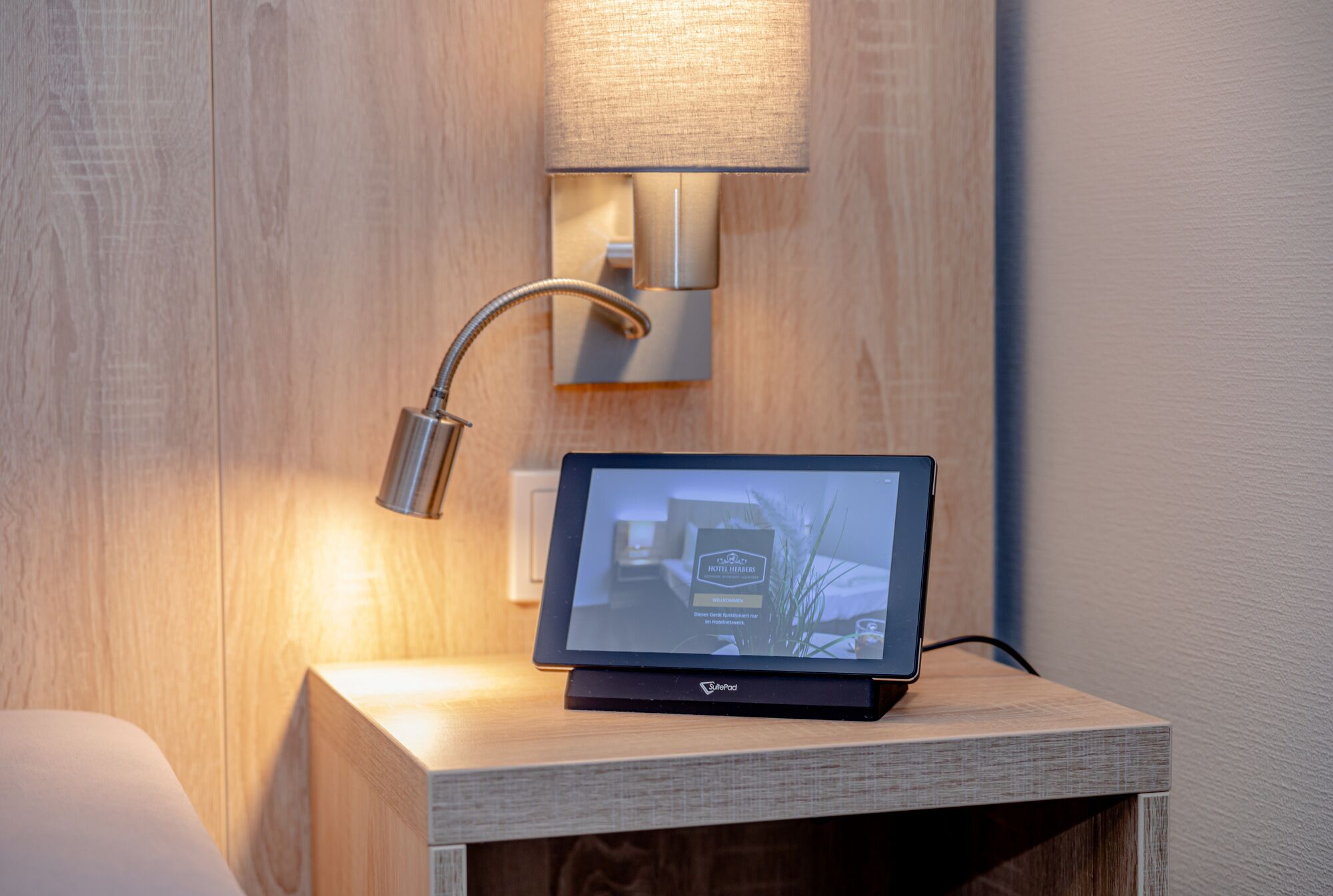 A tablet on a wooden nightstand with a wall-mounted lamp and a power outlet.