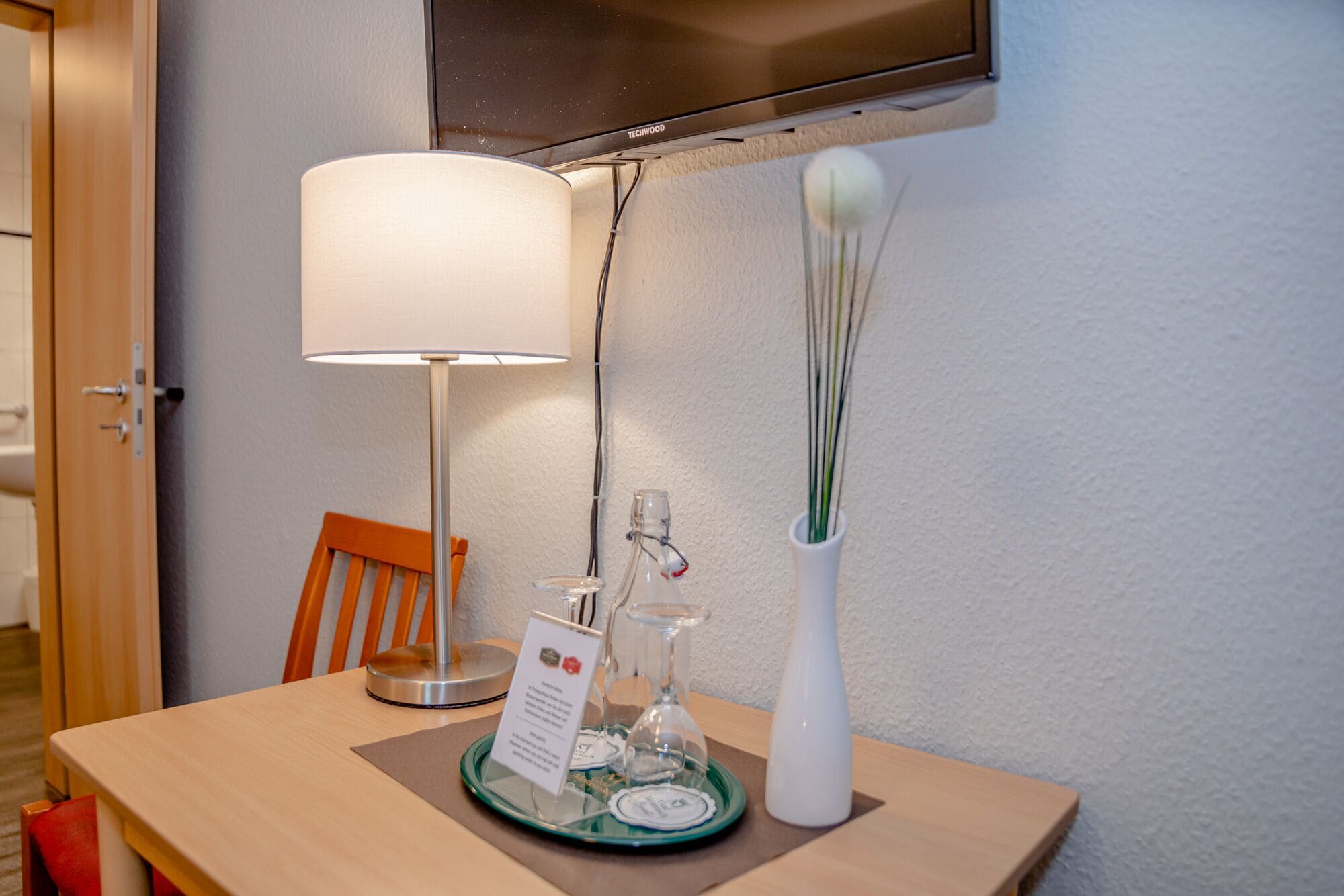 A hotel room with a wooden table, a lamp, a bottle, a vase, and a sign.