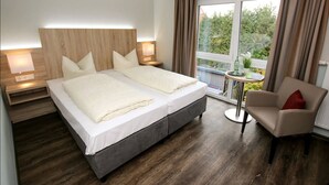 A double bed with a wooden headboard, two bedside tables with lamps, a small round table with a plant, a beige armchair, and a view of greenery through a window. - Hotel Herbers & RestaurantLeon (Sudbrookmerland)