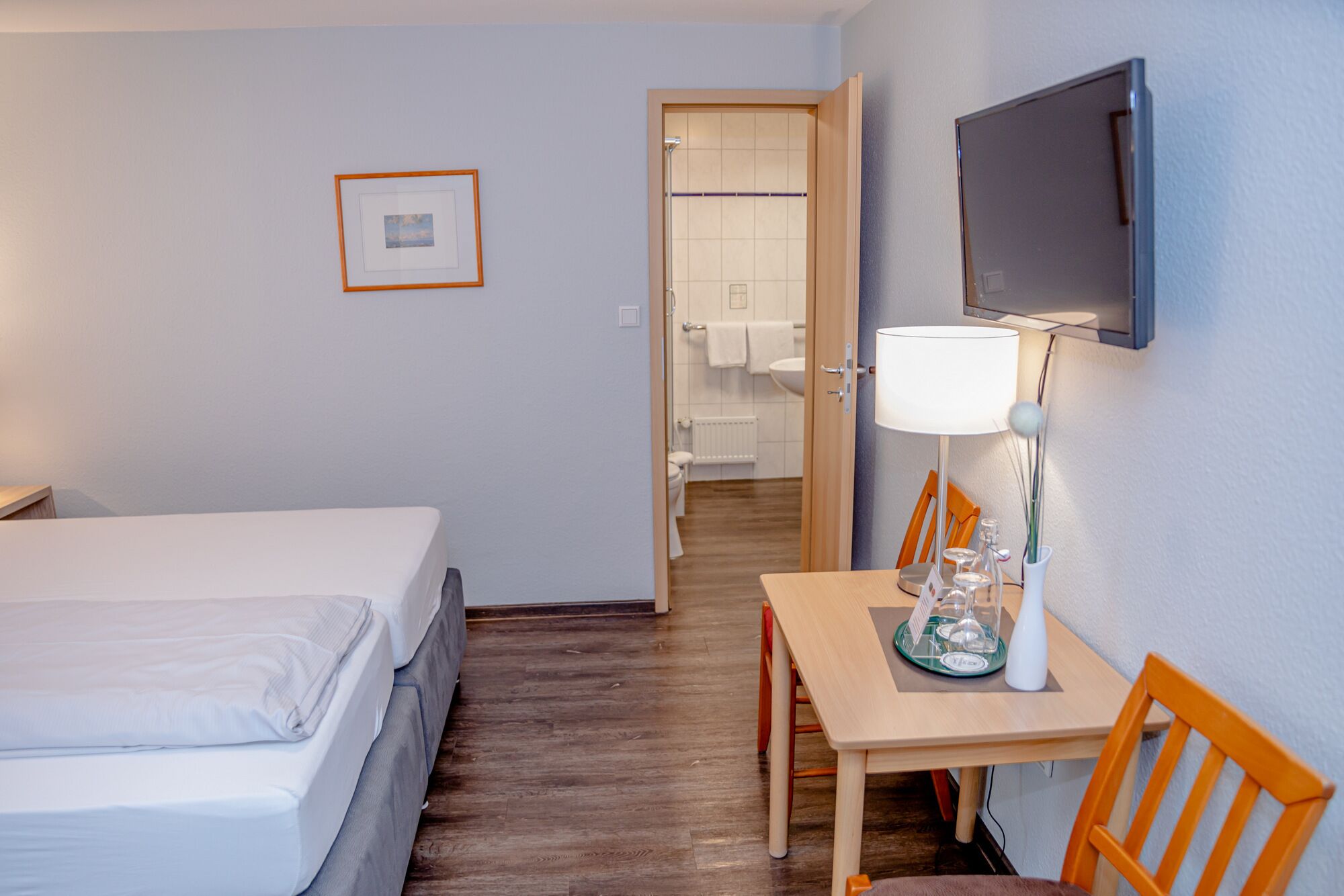 Una habitación de hotel con cama, escritorio con lámpara, televisor montado en la pared y un cuadro enmarcado.
