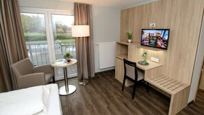 A hotel room with a bed, a desk, a chair, a TV, a small table with a lamp, a window with curtains, and a view of the outside. - Hotel Herbers & RestaurantLeon (Sudbrookmerland)