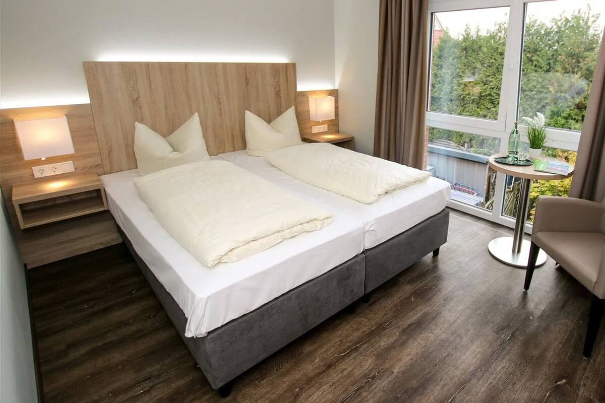 Een tweepersoonsbed met een houten hoofdeinde, een klein rond tafeltje met een plant, een stoel en uitzicht op groen buiten het raam.