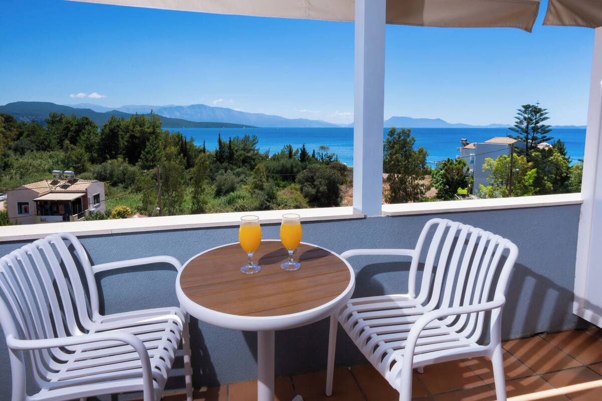 Un balcon avec deux chaises blanches et une petite table, deux verres de jus d’orange et une vue sur la mer et les montagnes.