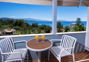A balcony with two white chairs and a small table, two glasses of orange juice, and a view of the sea and mountains.