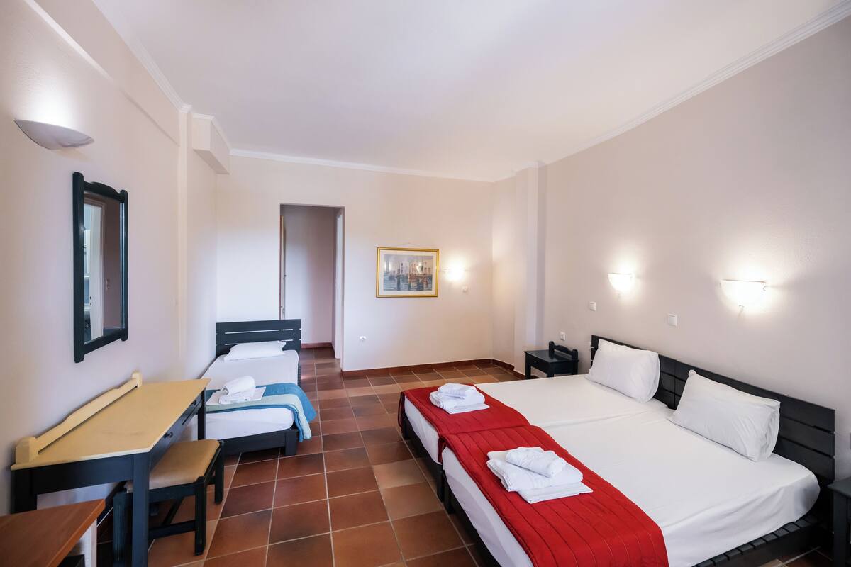 Une chambre d’hôtel avec deux lits, un bureau et une chaise. Il y a un tableau au mur et une porte qui donne sur une autre pièce.