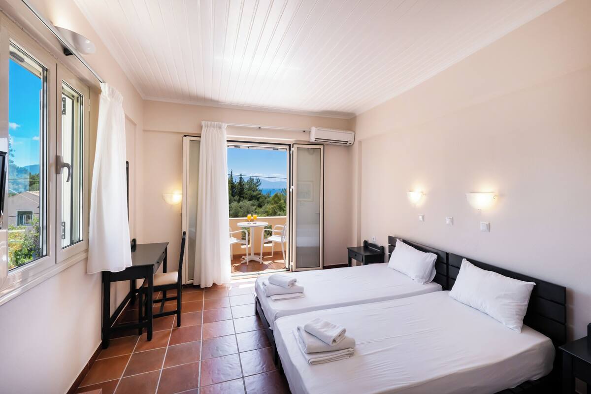 Une chambre d’hôtel avec deux lits, une petite table et une chaise. Elle dispose d’un balcon avec vue sur la mer et les montagnes.