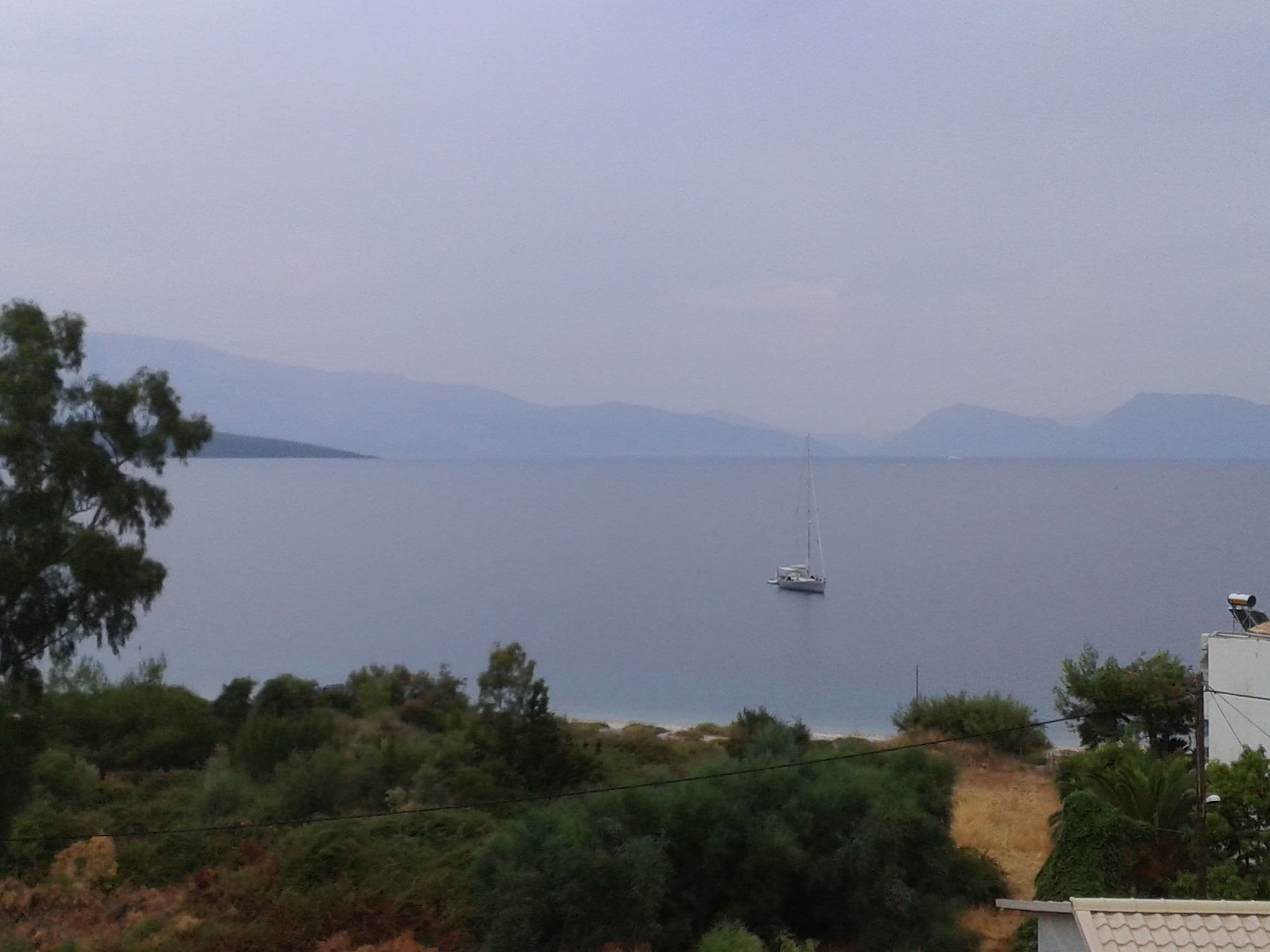 A view of a calm sea with a sailboat, surrounded by hills and vegetation.