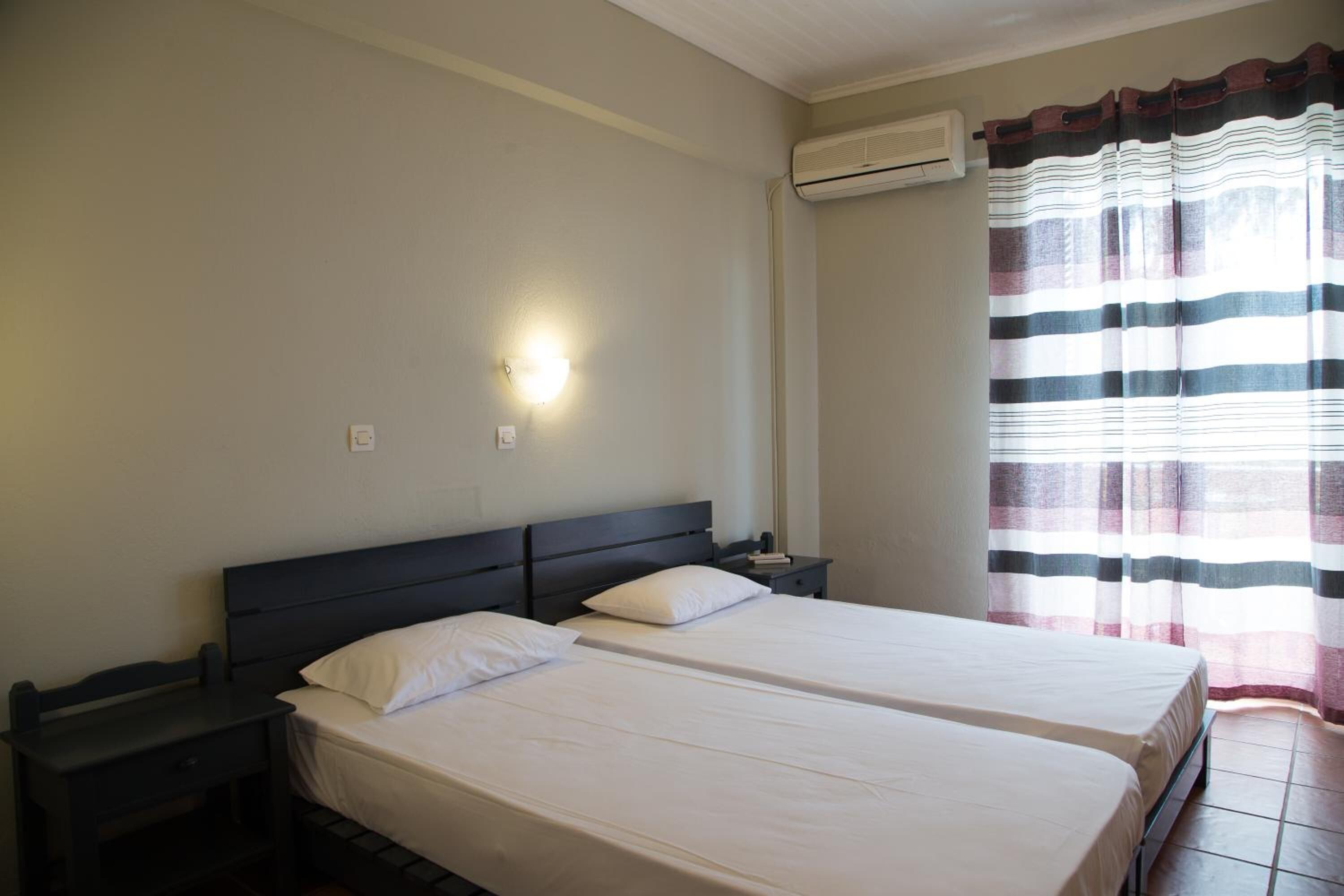 A room with two beds, a nightstand, a wall-mounted light, and a window with striped curtains.