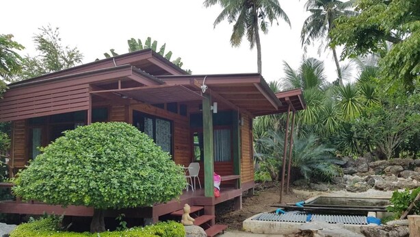 A wooden cabin with a porch, surrounded by lush greenery and palm trees. - Nung Ni Bangkonthi (Bang Khonthi)