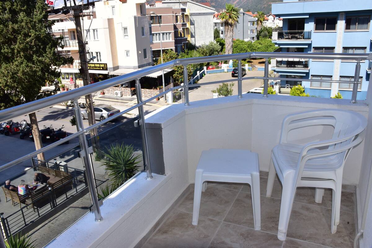 A balcony with a white plastic chair, a metal railing, and a view of a street with parked cars and buildings.