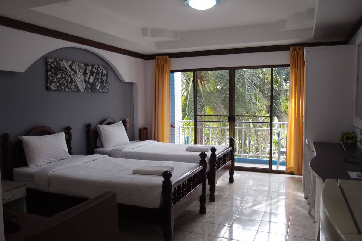 A hotel room with two beds, a desk, a chair, a balcony with a view of palm trees, and a ceiling light.