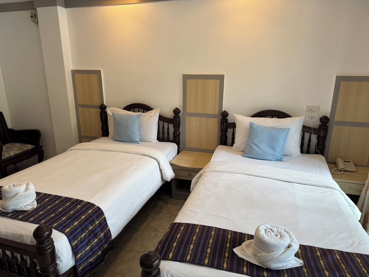 A hotel room with two single beds, each with a wooden headboard and white bedding. There are two blue pillows on each bed, and towels are neatly folded on the beds.