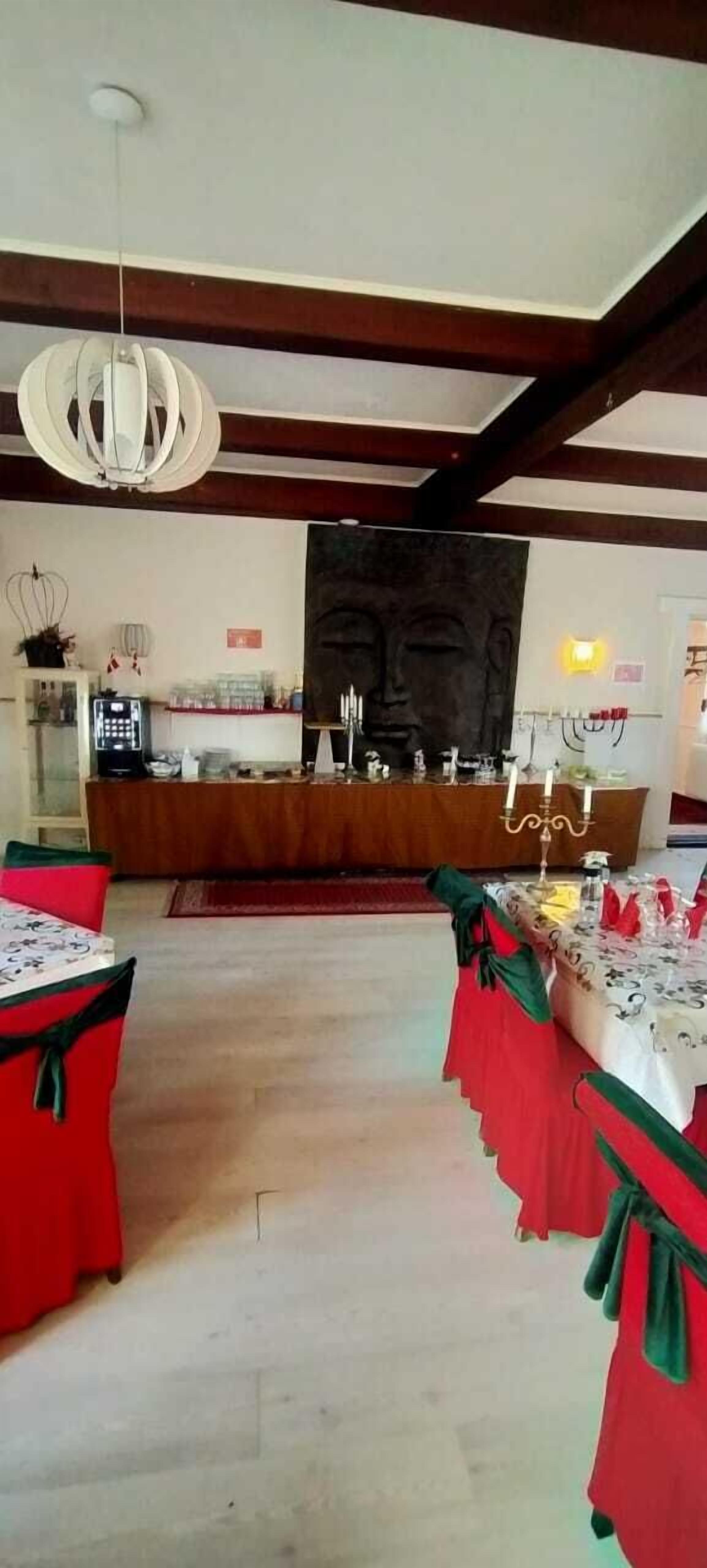 A spacious hall with a high ceiling and exposed beams, featuring a central altar with candles and a large religious painting. Tables with red and green tablecloths are set up for a gathering.