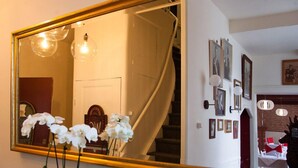 A room with a large mirror reflecting a staircase, a wall with framed pictures, and a vase with white flowers.