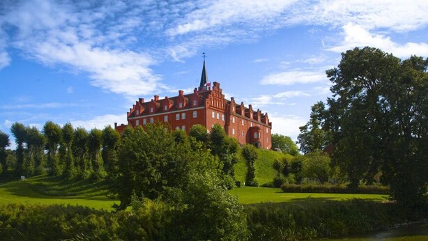 A large, historic red building with a prominent tower, surrounded by lush green trees and a well-maintained lawn.