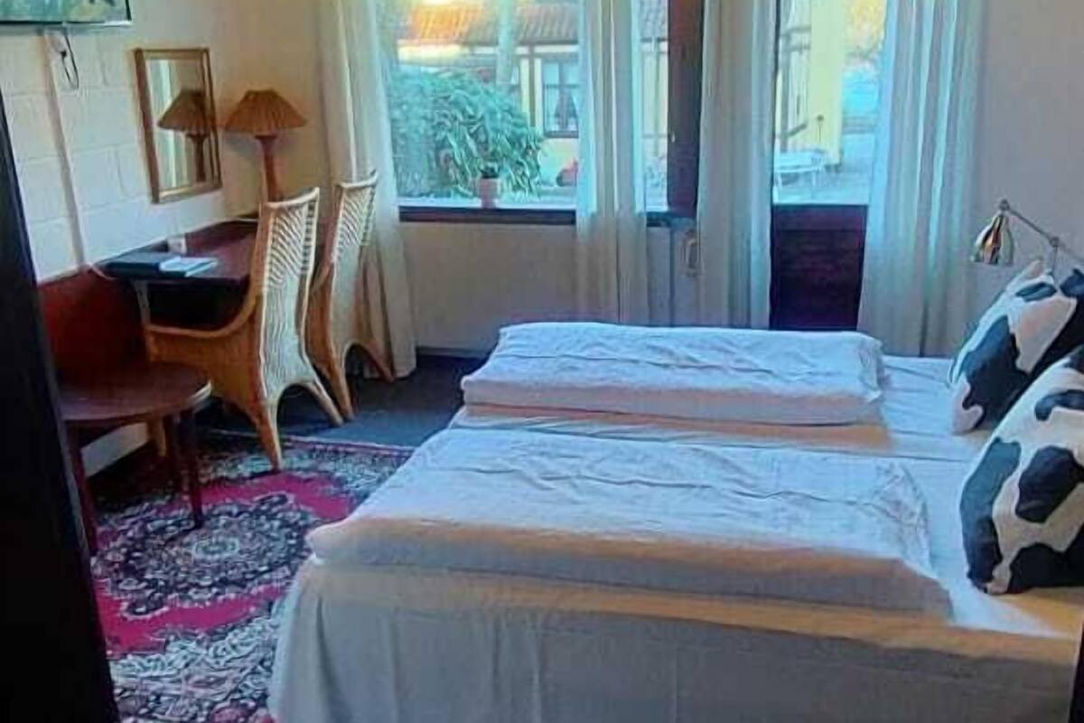 A hotel room with two beds, a desk, and a chair. There is a window with curtains, a chandelier, and a view of the outside.