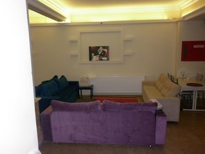 Una sala de estar con un sofá morado, un sofá azul, un sofá beige y una mesa de comedor con sillas. Hay un cuadro enmarcado en la pared y un detector de incendios rojo.