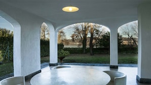 A modern outdoor dining area with a round table and four white chairs, surrounded by white pillars and a view of the outdoors. - Ritratto sul Lago (Dormelletto)