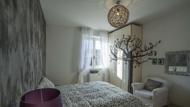 A bedroom with a bed, a chair, a lamp, a tree mural, and a window with curtains. - Ritratto sul Lago (Dormelletto)