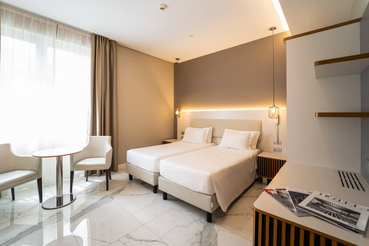 Camera d'albergo moderna con due letti, un piccolo tavolo, una sedia e una scrivania con una lampada.