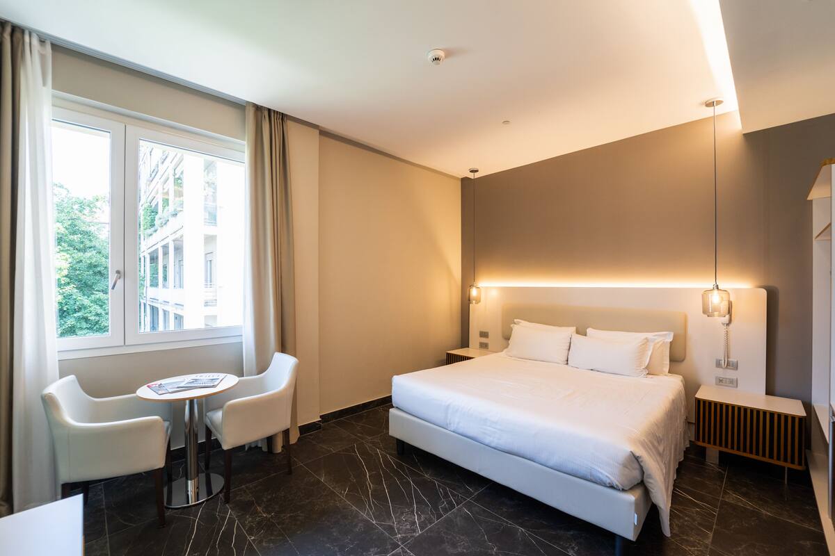 Una camera d'albergo con un letto grande, una scrivania, due sedie, una finestra con tende e vista sugli edifici.