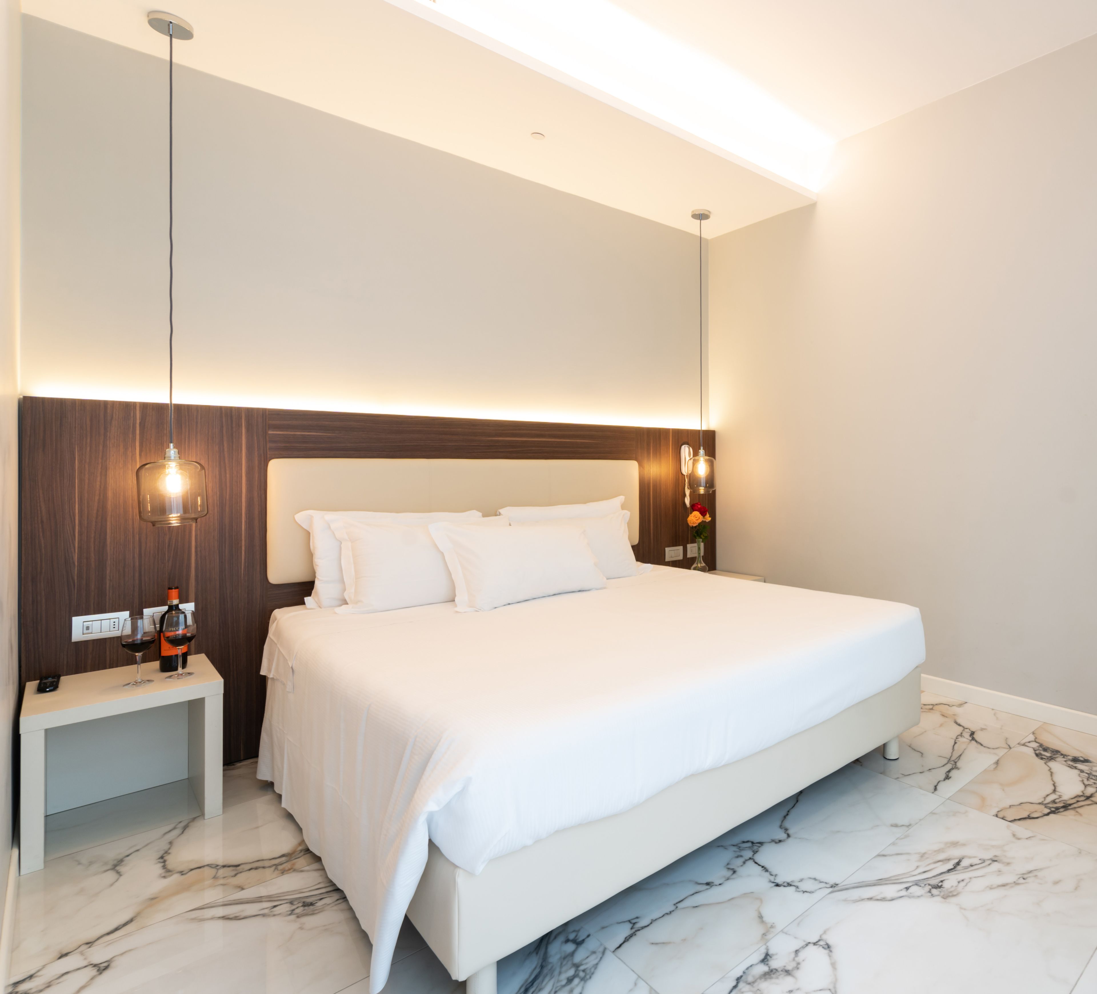 A modern hotel room with a large bed, marble flooring, a bedside table with a wine bottle, and a nightstand.