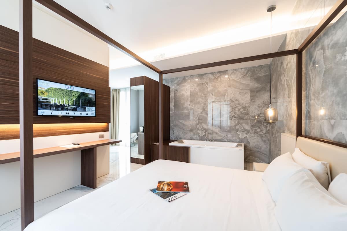 A modern hotel room with a large bed, a flat-screen TV mounted on a wooden panel, a marble accent wall, and a glass shower enclosure.