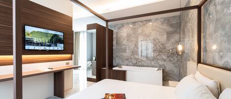 A modern hotel room with a large bed, a flat-screen TV mounted on a wooden panel, a marble accent wall, and a glass shower enclosure.