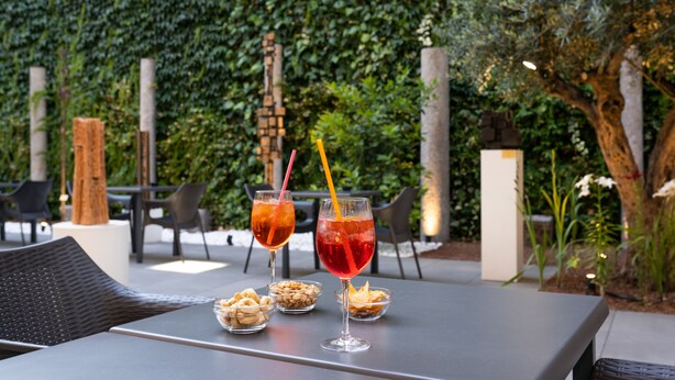 A table with two drinks and snacks, surrounded by chairs and a green hedge in the background. - Hotel Milano Castello (Milan)