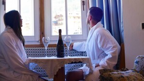 Une pièce aux murs en bois, une table recouverte d’une nappe en dentelle, deux fenêtres en arc de cercle et une bouteille de vin.