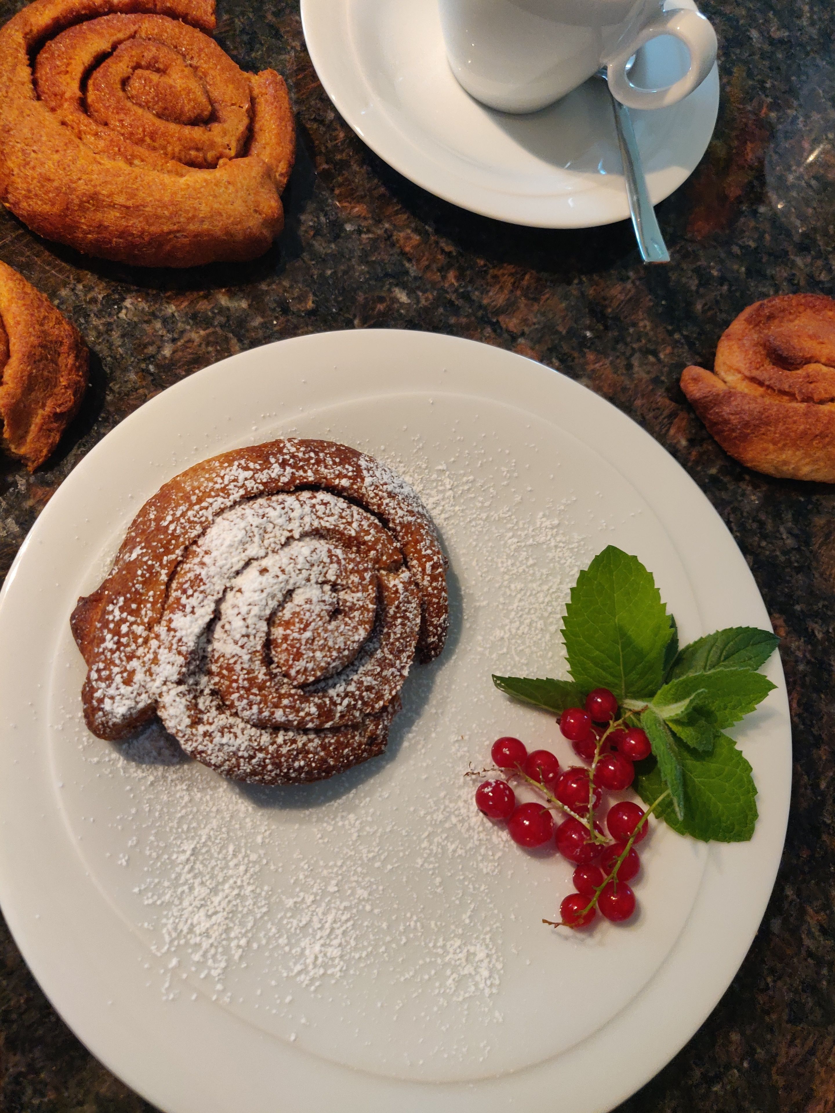 A plate with a rolled pastry dusted with powdered sugar, accompanied by a sprig of mint and red berries, and a cup and saucer in the background.