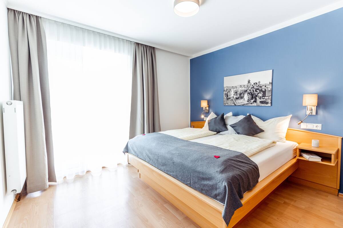 Une chambre d’hôtel moderne avec un lit en bois, une grande fenêtre avec des rideaux, un mur d’accent bleu et un tableau encadré.