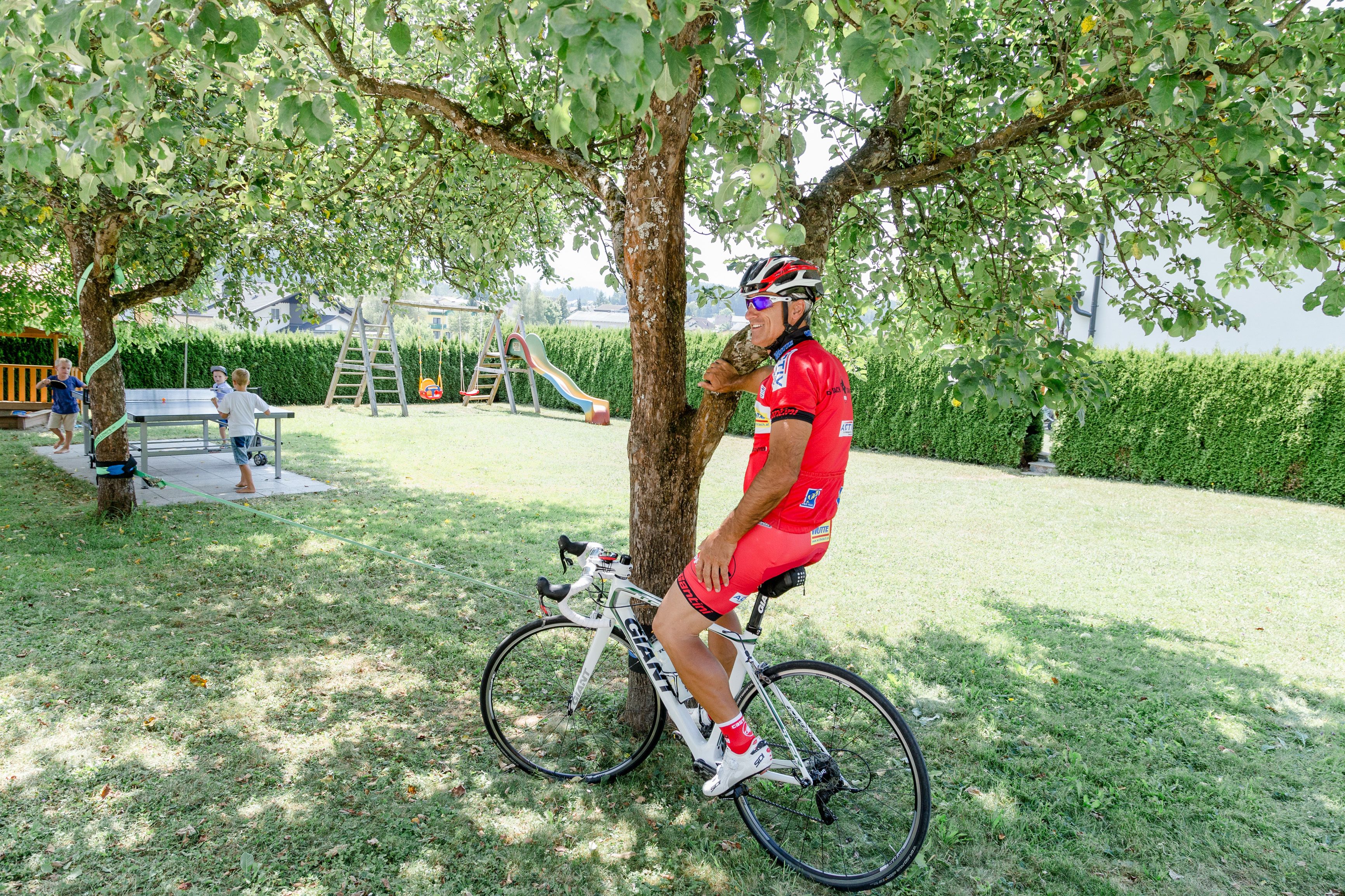 A person on a bicycle in a grassy area with a tree, a slide, and a swing set in the background.