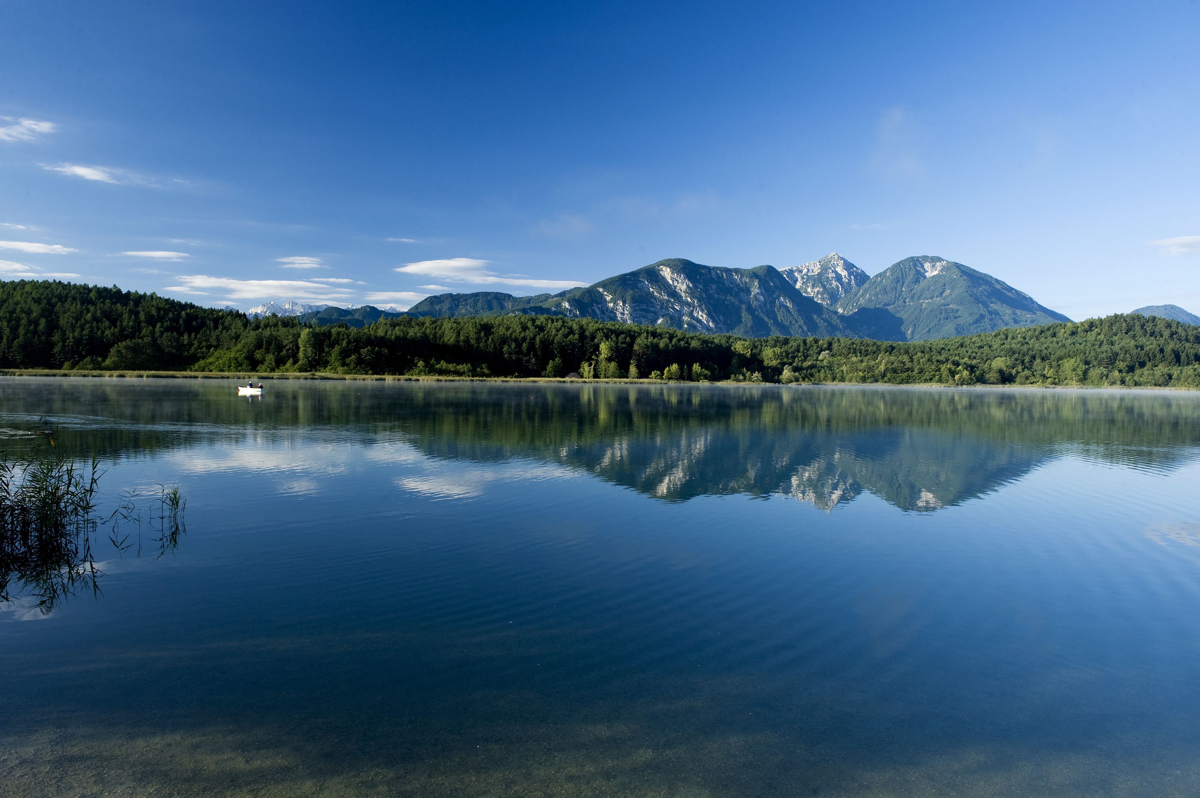 Ein ruhiger See mit einem Boot, umgeben von bewaldeten Bergen unter blauem Himmel.