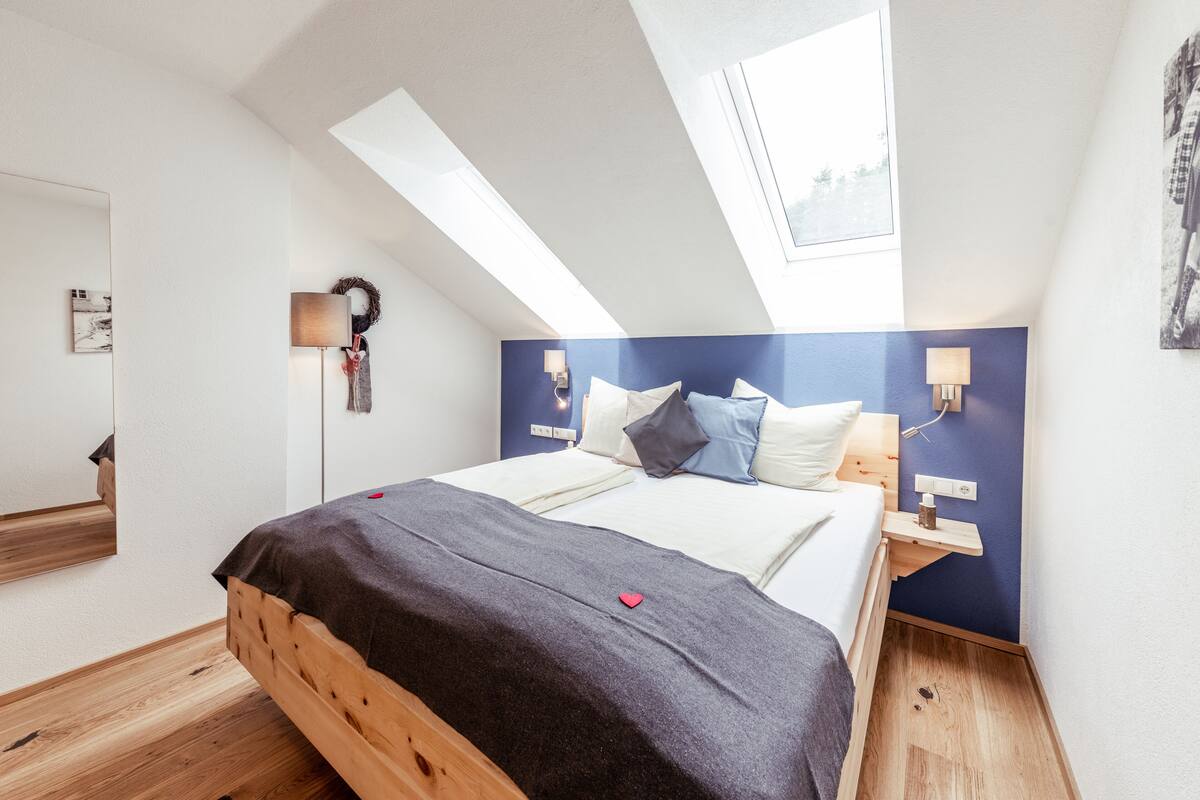 Une chambre avec un puits de lumière, un lit recouvert d’une couverture foncée, une table de chevet avec une lampe et un luminaire fixé au mur.