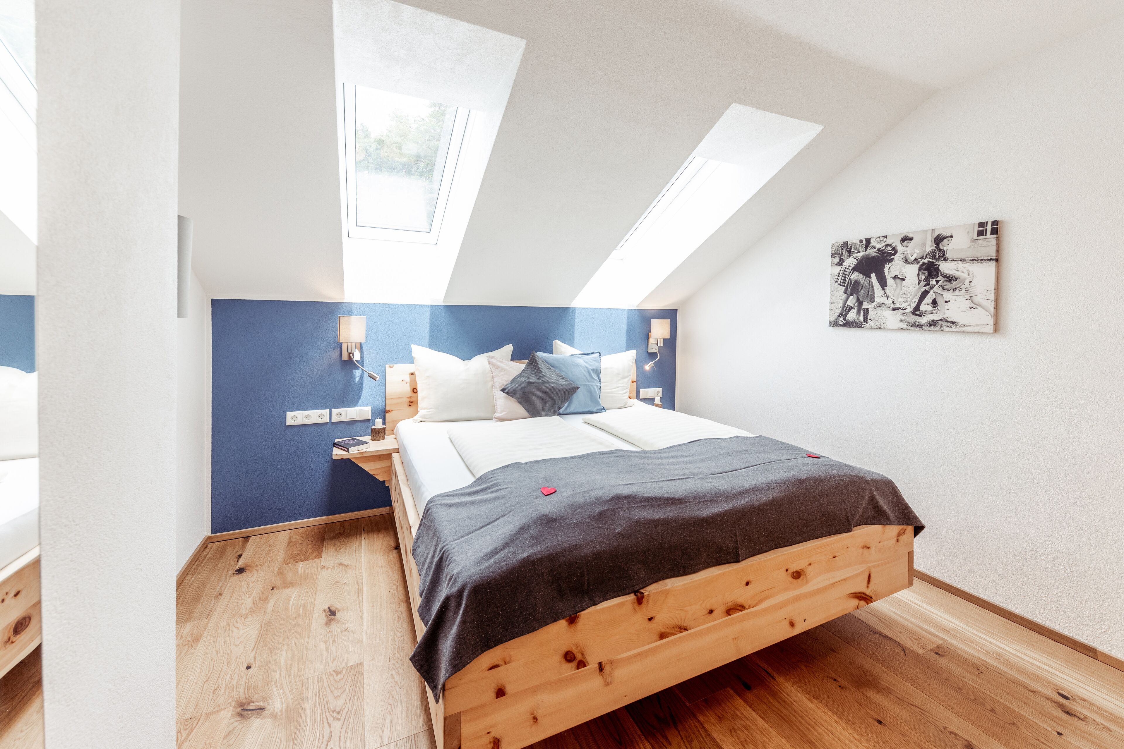 A bedroom with a sloped ceiling, skylights, a bed with a wooden frame, a blue accent wall, and a wall-mounted light fixture.