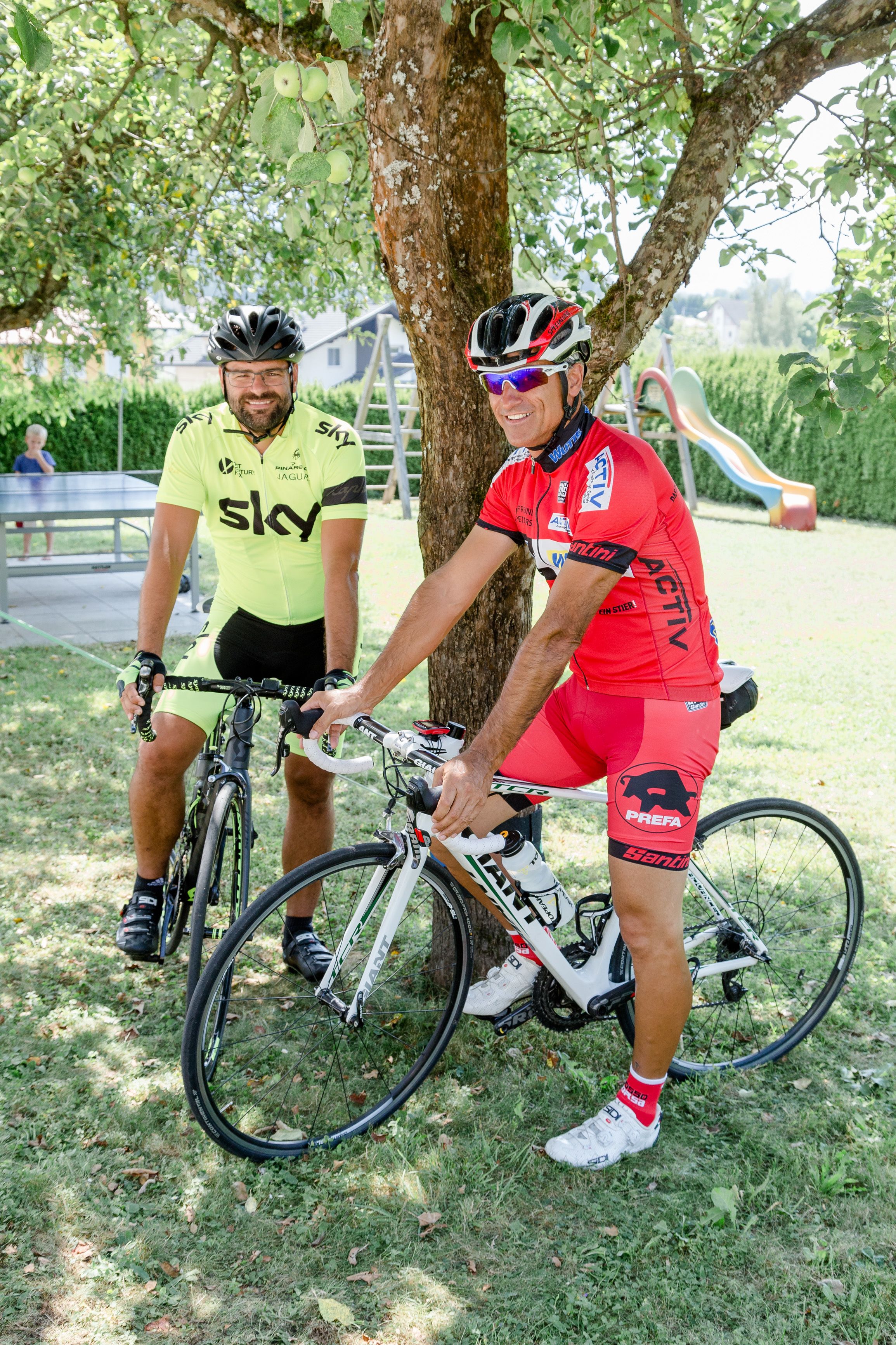 Two cyclists sitting on bicycles under a tree in a park.