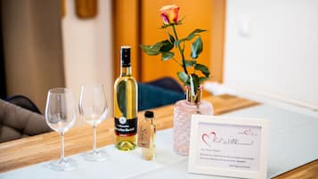 Una mesa con copas de vino, una botella de vino, un frasco de perfume, un florero rosa con una rosa y una tarjeta enmarcada.