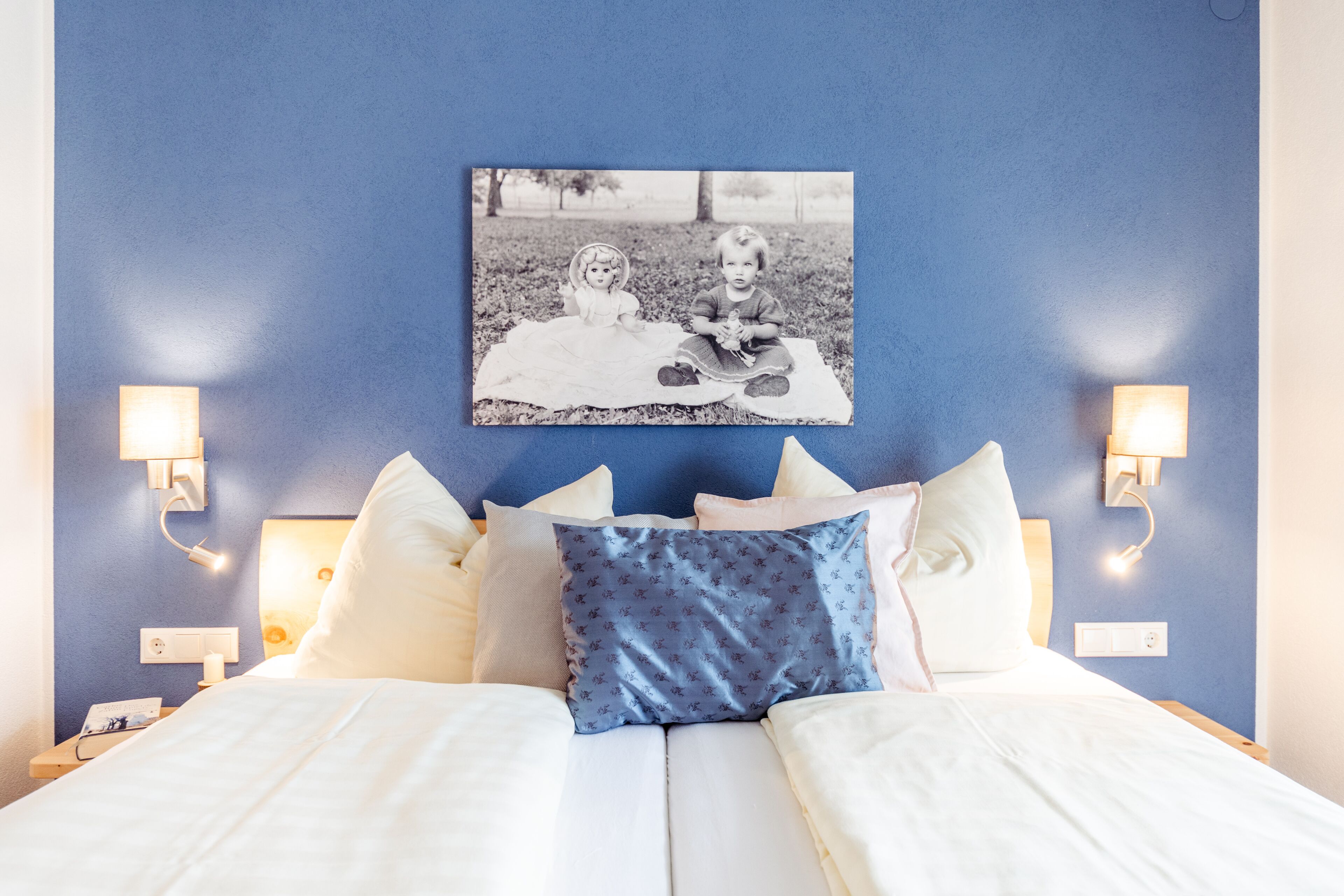 A neatly made bed with white linens and a blue patterned pillow, a black and white photo of two children on a blanket, and wall-mounted lamps on either side of the bed.
