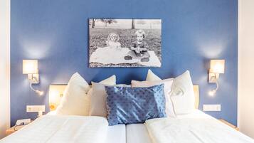 A neatly made bed with white linens and a blue patterned pillow, a black and white photo of two children on a blanket, and wall-mounted lamps on either side of the bed.