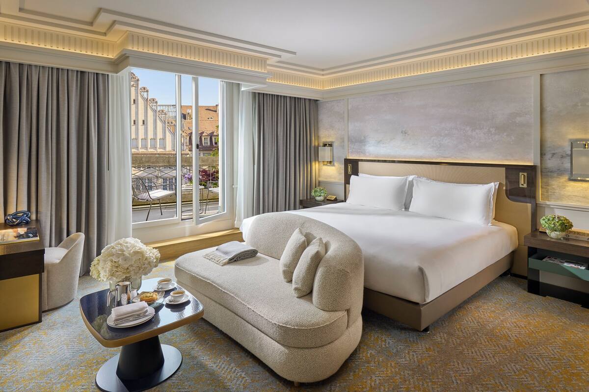 A hotel room with a large bed, a sofa, a desk with a vase of flowers, and a view of the city.