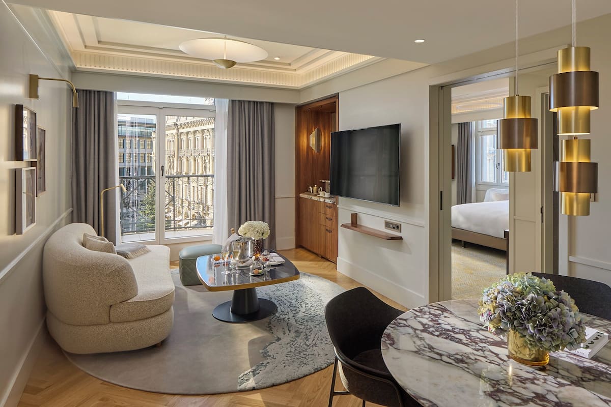 A modern hotel room with a living area, a dining table, a sofa, a TV, and a balcony with a city view.